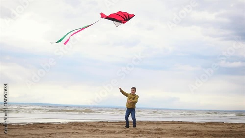 Boy in jacket playing with kite on the beach. Windy cold weather and waves in the ocean