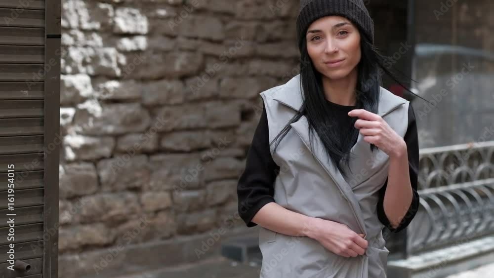 Stylish Fashion Girl Smile, With Long Black Hair, Walking in the Street, on Background Brick Wall, in Grey Cloak and Black Hat Stylish Fashion Girl Smile, With Long Black Hair, Walking in the Street, on Background Brick Wall, in Grey Cloak and Black Hat