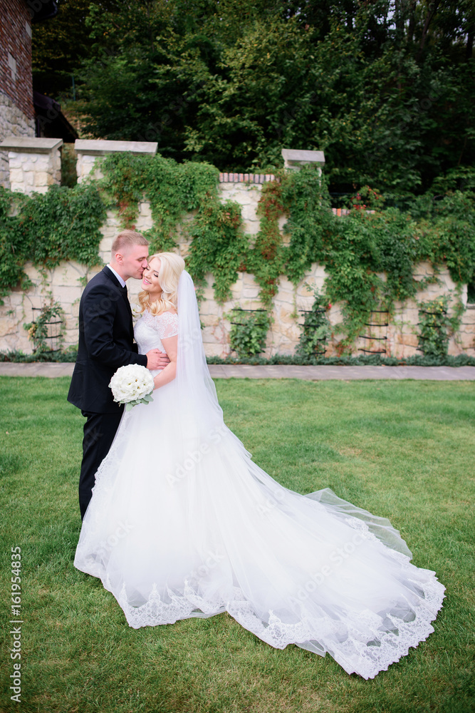 Groom in black suit and classy blonde bride stand on the green backyard Groom in black suit and classy blonde bride stand on the green backyard