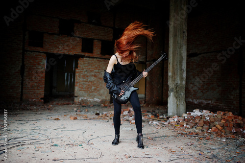 Red haired punk girl wear on black with bass guitar at abadoned place. Portrait of gothic woman musician.