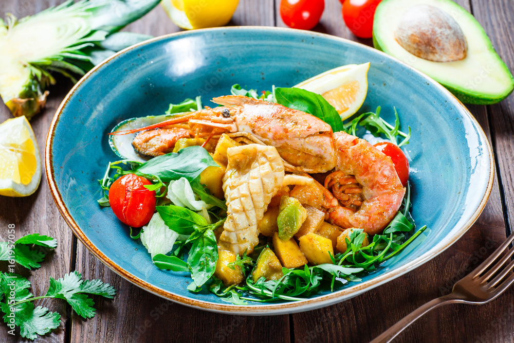 Warm salad with seafood, langoustine, mussels, shrimps, squid, scallops, mango, pineapple, avocado, arugula and basil on wooden background close up. Mediterranean food. Top view Warm salad with seafood, langoustine, mussels, shrimps, squid, scallops, mango, pineapple, avocado, arugula and basil on wooden background close up. Mediterranean food. Top view