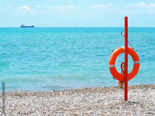 Red and orange lifebuoy on a pebble beach. Life buoy on the background of the blue sea.