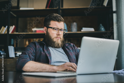 Portrait of young casual bearded man working on laptop at home