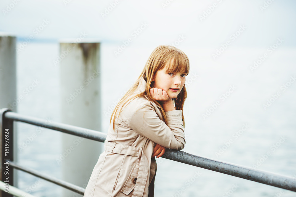 Outdoor portrait of adorable 9 -10 year old kid girl wearing modern beige trench coat, posing in a port on a cold day