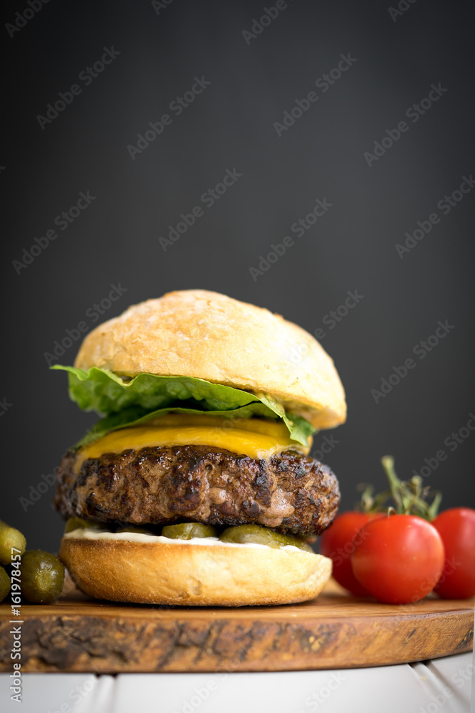 large thick grilled beef minced burger patty with toasted bun melted cheddar cheese green fresh salad cherry tomato and pickle cucumber on rustic wooden board and white table