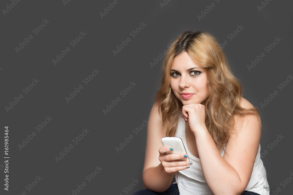 Studio shot of attractive long haired teenage girl holding her smart phone and looking at the camera. All is on the gray background. All potential trademarks and buttons are removed. Studio shot of attractive long haired teenage girl holding her smart phone and looking at the camera. All is on the gray background. All potential trademarks and buttons are removed.