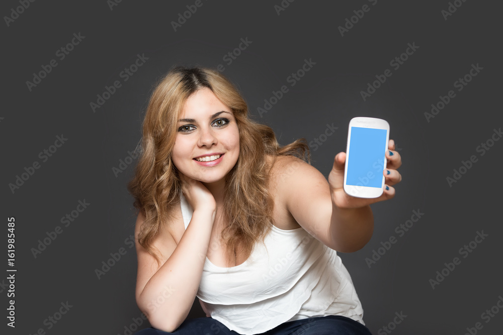 Long haired smiling teenage girl showing smart phone with blank touchscreen. The girl is holding her head by right hand. All is on the gray background. Long haired smiling teenage girl showing smart phone with blank touchscreen. The girl is holding her head by right hand. All is on the gray background.