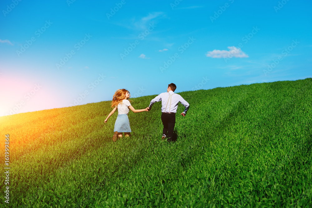 Young happy lovers running on meadow with green grass and blue sky, From the back Young happy lovers running on meadow with green grass and blue sky, From the back