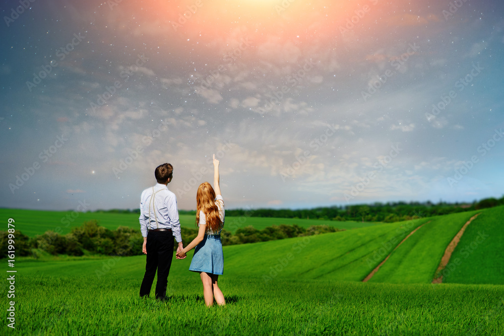 Young couple standing on green field, woman showing her hand at the stars