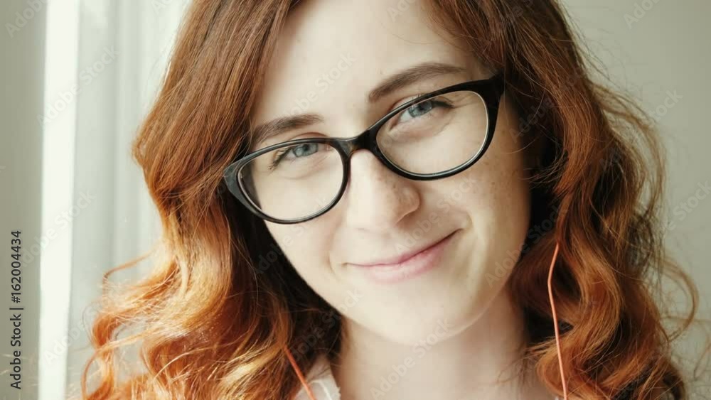 Beautiful redhead girl. Close up. Pretty girl looking at camera and smiling. Young woman in glasses listening to music on headphones and texting. Woman using mobile cell smart phone Beautiful redhead girl. Close up. Pretty girl looking at camera and smiling. Young woman in glasses listening to music on headphones and texting. Woman using mobile cell smart phone