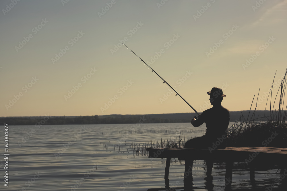 A fisherman on the river bank, sits on a wooden bridge and catches fish.