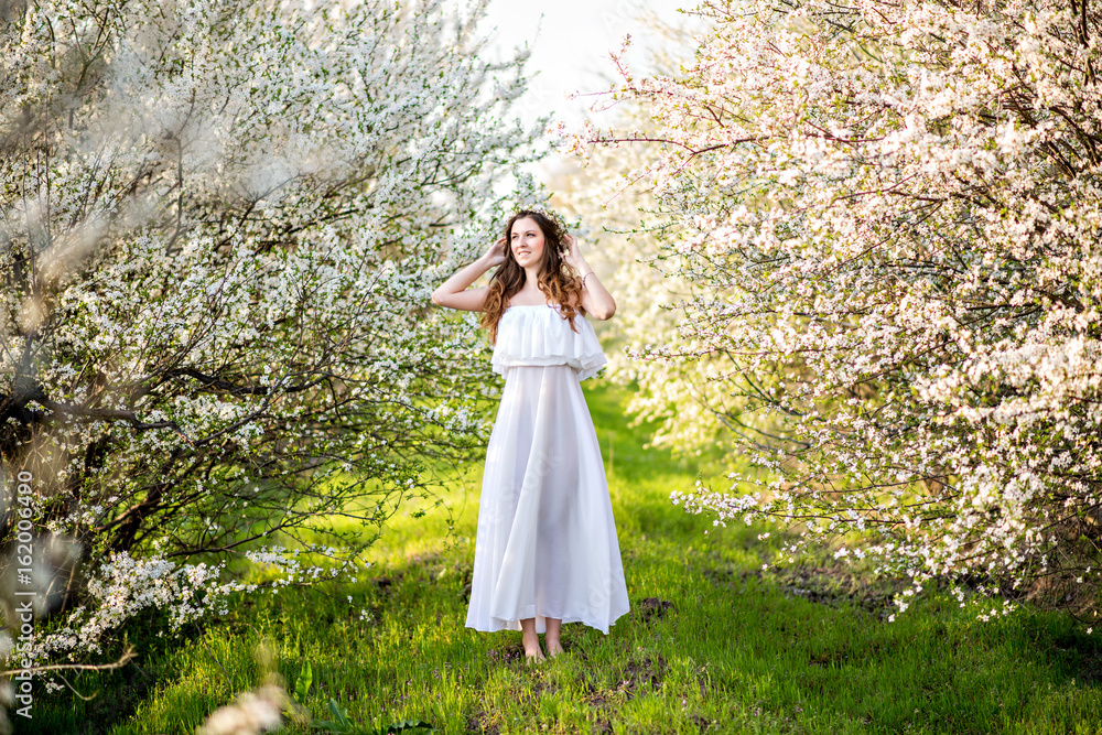 Beautiful young girl in white dress and wreath of flowers on head in blooming gardens Beautiful young girl in white dress and wreath of flowers on head in blooming gardens