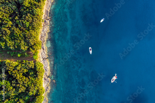 Coastal area with three boats on blue clear water and forest on land - aerial view taken by drone