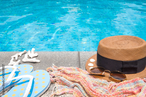 Concept of holiday tropical on summer, Accessories blue slippers, rainbow scarf, straw hat, shell, coral and sunglasses on border of a swimming pool.