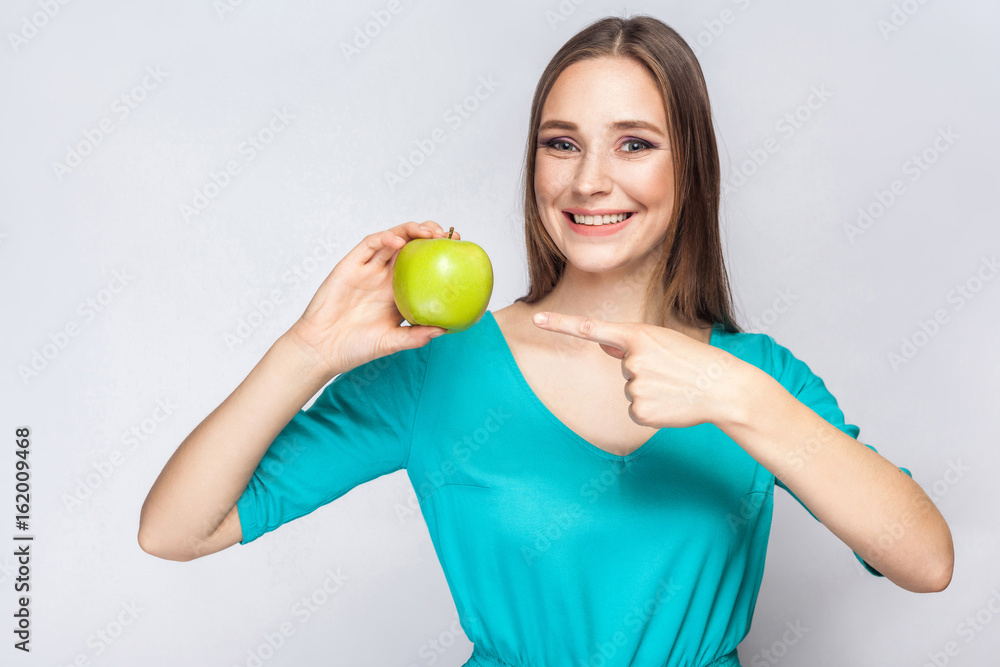 Young beautiful woman with freckles and green dress holding apple and pointing with finger. studio shot, isolated on light gray background. Young beautiful woman with freckles and green dress holding apple and pointing with finger. studio shot, isolated on light gray background.