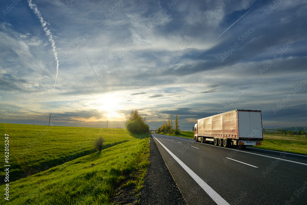 Red truck driving on the asphalt road between fields and meadows in rural landscape at sunset