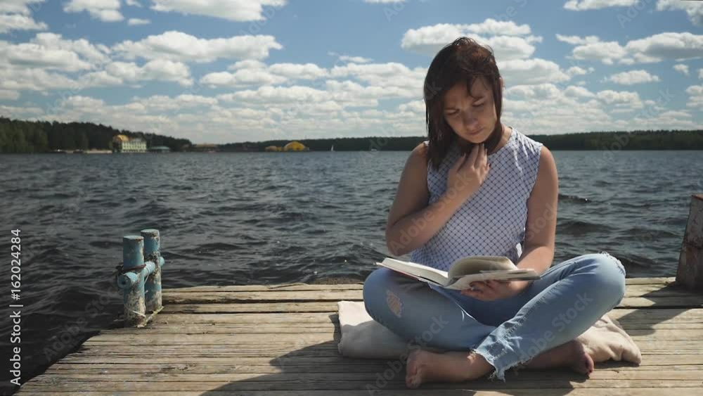 Young brunette sitting on old wooden pier in the lotus pose and reading book on background of lake and forest Young brunette sitting on old wooden pier in the lotus pose and reading book on background of lake and forest