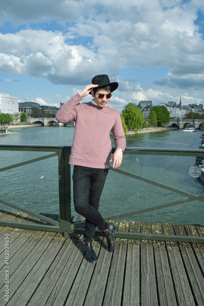 Paris,France -April 2,2017: Young Good looking Asian man in pink shirt and black hat on Pont des Arts bridge in Paris. Paris,France -April 2,2017: Young Good looking Asian man in pink shirt and black hat on Pont des Arts bridge in Paris.