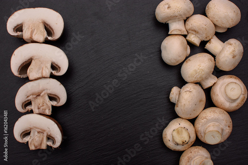 White mushrooms champignons whole and sliced lie on a stone black slate board, background