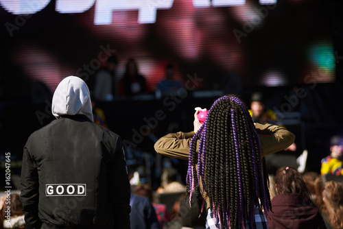 Rear view of stylish couple partying at rock concert, electronic music festival, dance floor or disco club. Crowd of young people standing in front of stage, listening to music band performance photo