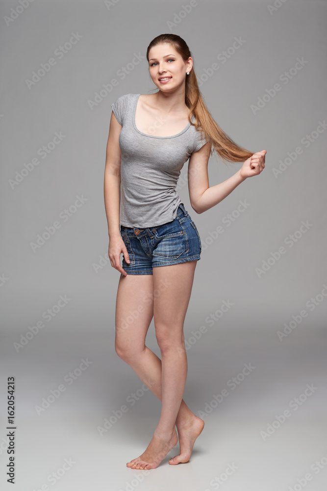 Beautiful barefooted full length female in denim shorts and grey tshirt posing over grey background holding playing with long hair Beautiful barefooted full length female in denim shorts and grey tshirt posing over grey background holding playing with long hair