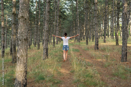 A woman walks through pine forest with open hands. Rear view.