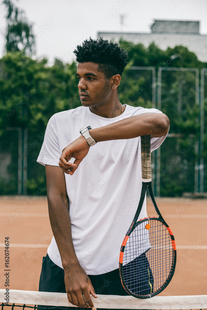 Concentrated young man holds his hand on a tennis racket. Portrait of serious african man looking away.