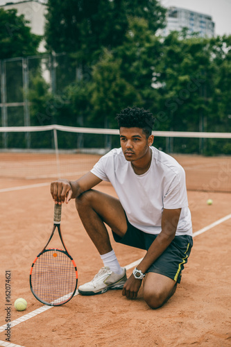 Young man hanging out in the tennis courts sitting on ground. African man resting and chilling on the break time.