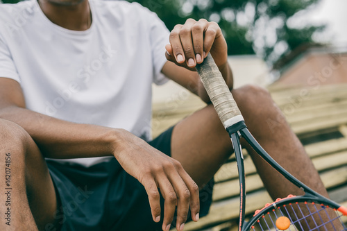 Close-up of a tennis player holding a racket sitting on a bench. The sportsman has a rest from below.