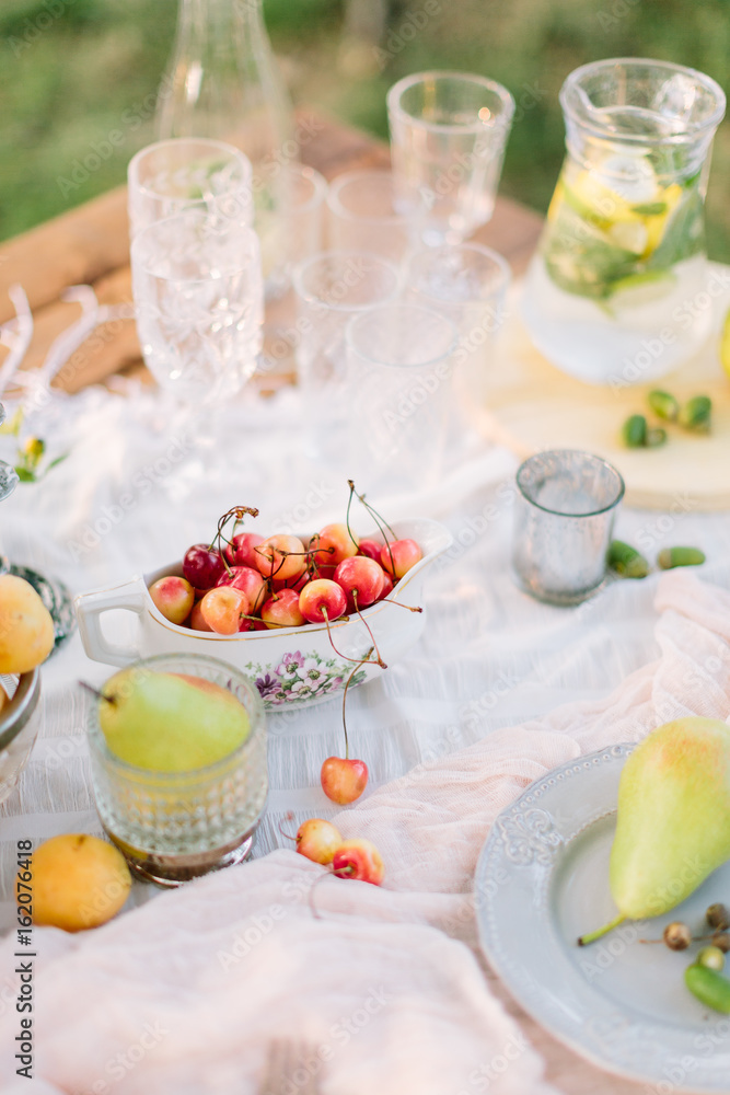 picnic, food, summer, holiday concept - close-up on wooden festive table with white tablecloth, dish with cherries, served with glass cups, goblets, glasses, fruit plates, pitcher of lemonade. picnic, food, summer, holiday concept - close-up on wooden festive table with white tablecloth, dish with cherries, served with glass cups, goblets, glasses, fruit plates, pitcher of lemonade.