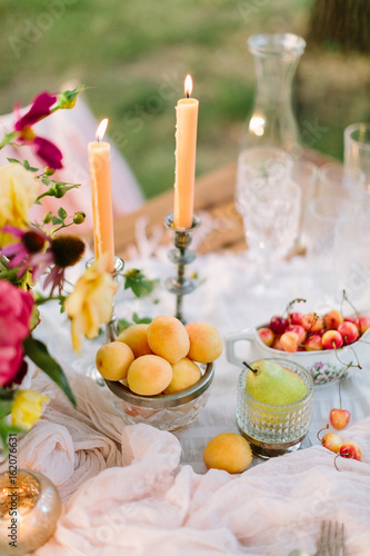 picnic, dating, wedding, valentines day, weekends concept - romantic table setting for newlyweds with flowers, burning candles, crystal glasses and such treat like apricots, pears and cherries