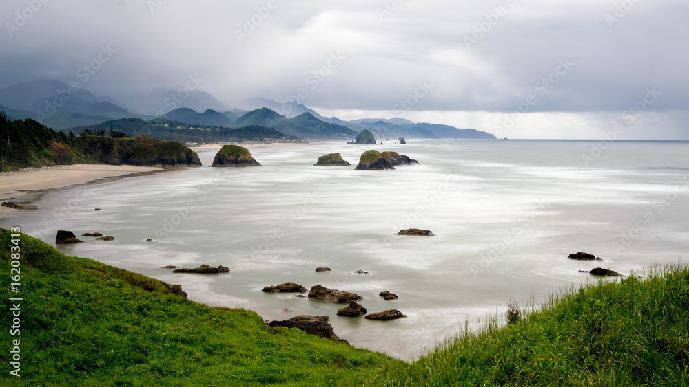 Long exposure of Cannon Beach in Oregon, USA featuring rocky outcrops in the ocean and mountains in the background on a cloudy sky day