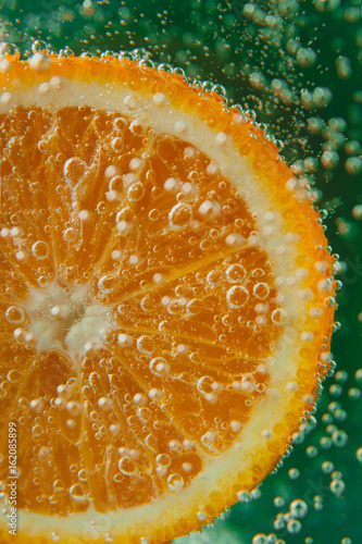Orange slice in water with bubbles on green background, macro photo