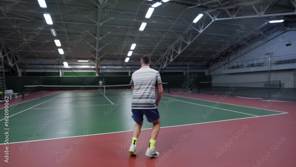 An adult man plays a match in tennis on a sports court, a person in a sporty form beats the ball with a racket in the gym An adult man plays a match in tennis on a sports court, a person in a sporty form beats the ball with a racket in the gym