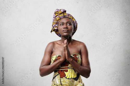 Praying African model with big dark eyes, smooth skin and stubby nose wearing traditional scarf and dress. Hopeful dark-skinned middle-aged woman keeping her beautiful hands together while worshiping