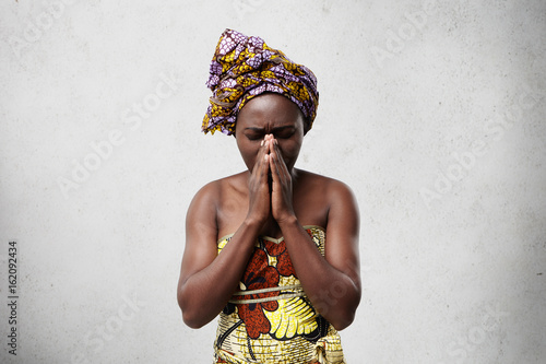 Billede på lærred Portrait of begging black woman in traditional clothes pressing her palms together closing her eyes imploring for good luck of her children