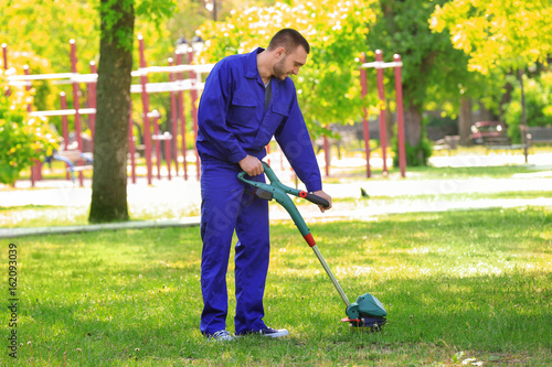 Young worker mowing lawn with grass trimmer outdoors on sunny day