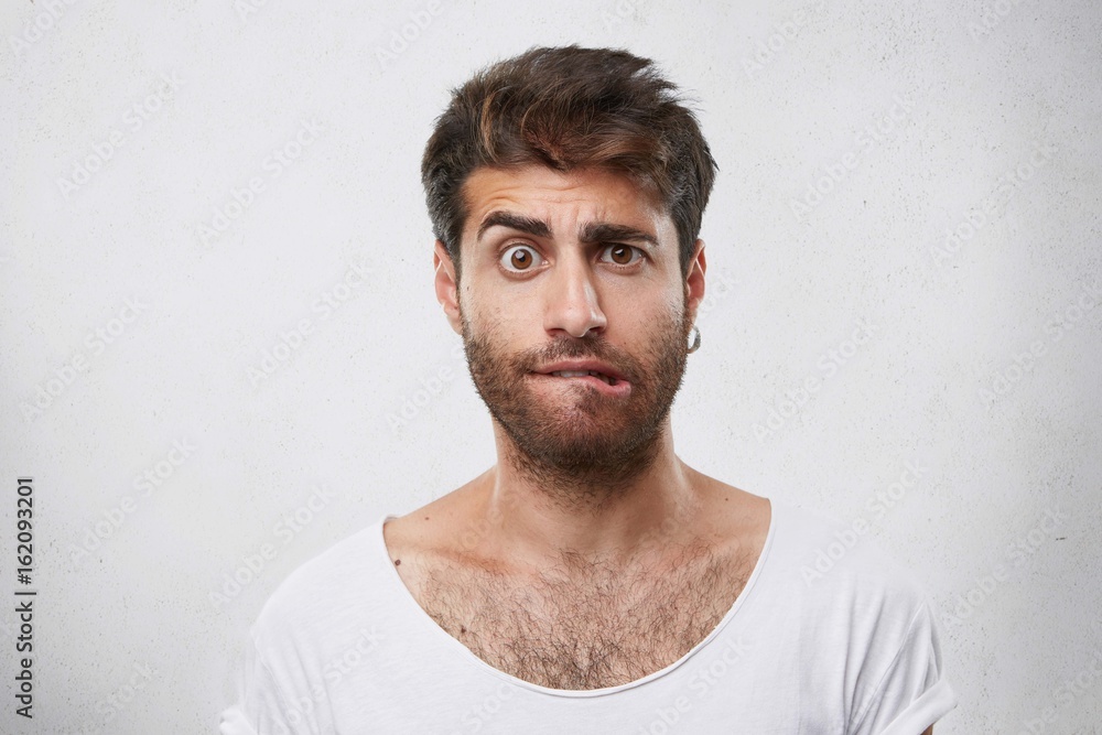 Fototapeta premium Nervous doubtful male with stylish hairstyle and beard frowning his eyebrow, biting lips having puzzled look going to make serious decision. Surprised macho man expressing his feelings and emotions