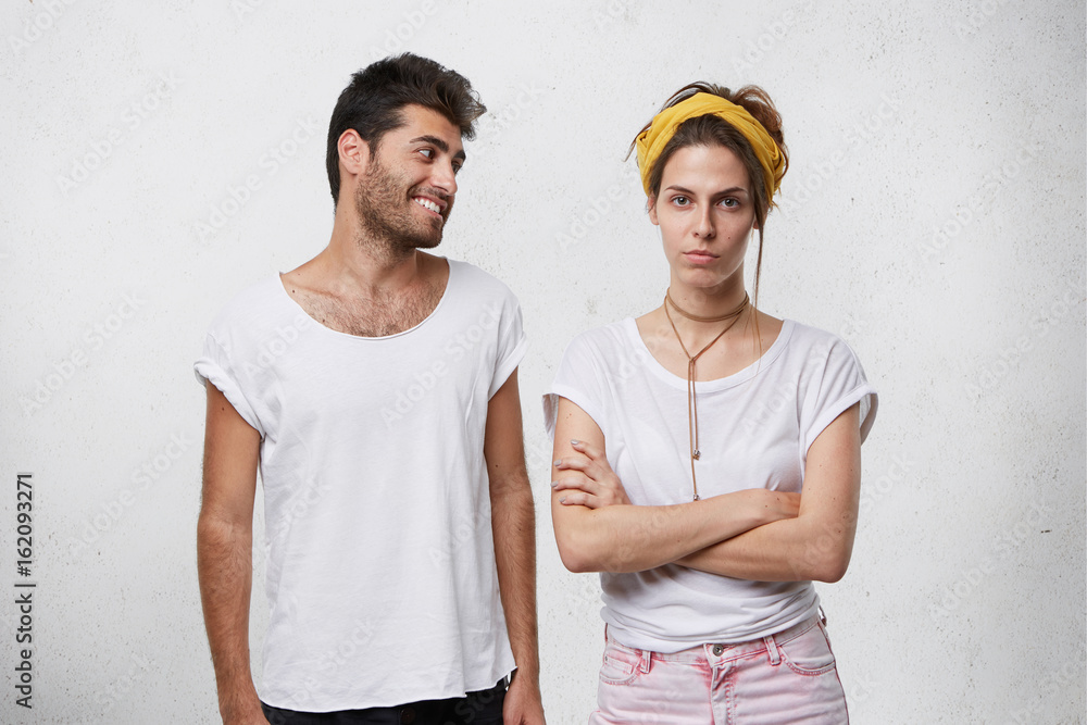 Studio shot of handsome positive bearded male in white t-shirt trying to convince or apologize to his angry upset girlfriend in yellow headband who is looking offended, keeping her arms crossed Studio shot of handsome positive bearded male in white t-shirt trying to convince or apologize to his angry upset girlfriend in yellow headband who is looking offended, keeping her arms crossed