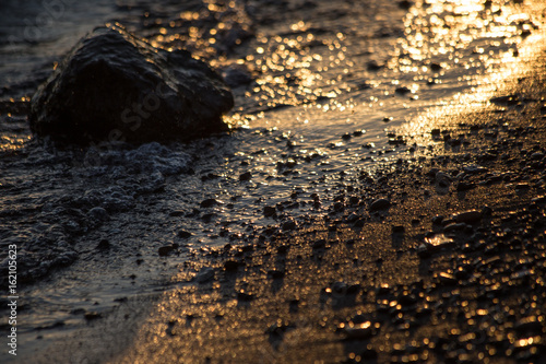 A close up of a shore at sunset, with light reflections on water and big and small stones