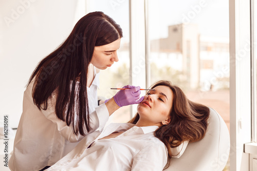 Female customer is lying on a white couch during correction and eyebrow styling. Young women tweezing her eyebrows in beauty salon.