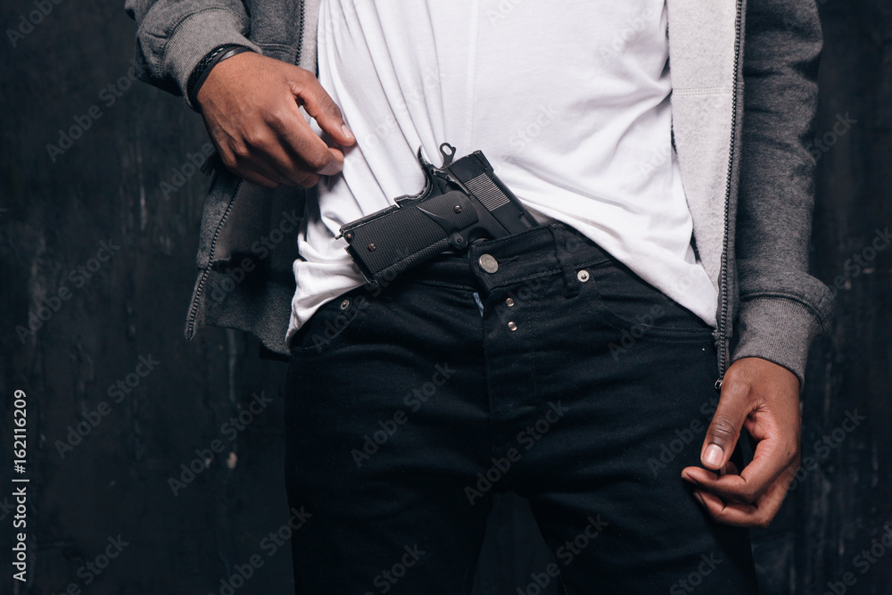 Unrecognizable black man threatens with a gun closeup studio shoot. Ghetto gangster with weapon on dark background. Outlaw, ghetto, murderer, robbery concept