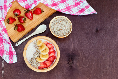 Smoothie bowl decorated with banana, strawberries and chia seeds with ingredients on a wooden table. Top view.