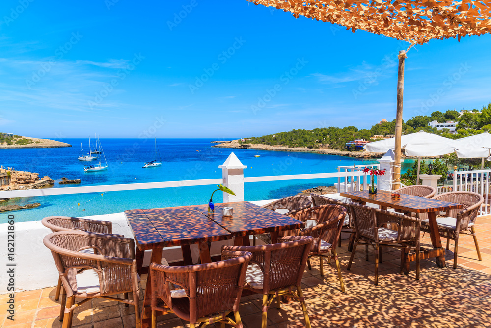 Tables with chairs on terrace of coastal restaurant in Cala Portinatx bay, Ibiza island, Spain.