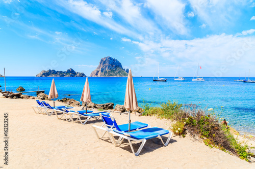 Sunbeds with umbrellas on Cala d'Hort beach with beautiful azure blue sea water and Es Vedra island in distance, Ibiza island, Spain