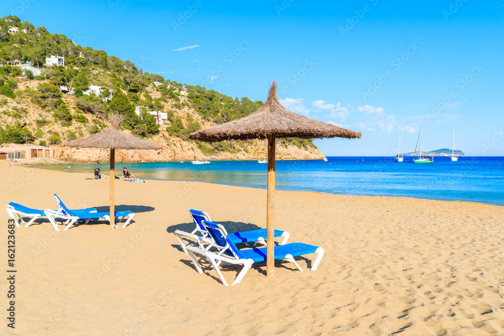 Sandy beach with umbrellas and sunbeds in Cala San Vicente bay on sunny summer day, Ibiza island, Spain