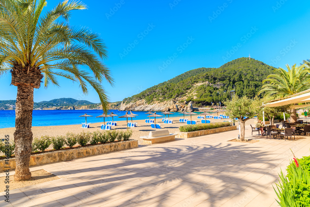 Naklejka premium Coastal promenade along sandy beach with restaurants and bars in Cala San Vicente bay on sunny summer day, Ibiza island, Spain