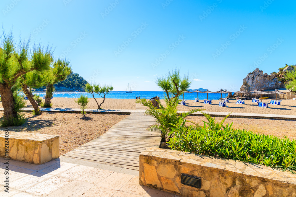 Walkway to sandy beach with umbrellas and sunbeds in Cala San Vicente bay on sunny summer day, Ibiza island, Spain