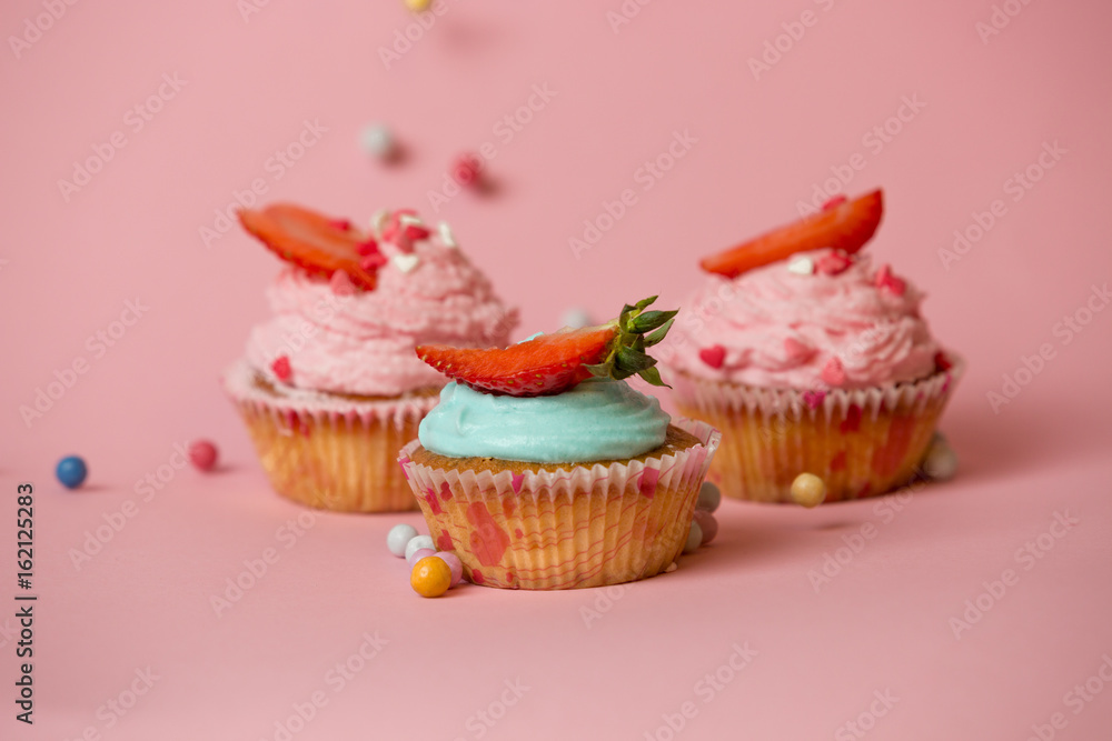 Three colorful cupcakes with strawberries on pink background with colorful candies Three colorful cupcakes with strawberries on pink background with colorful candies