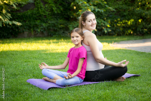 Mother and smiling daughter practicing yoga and sitting back to back at park